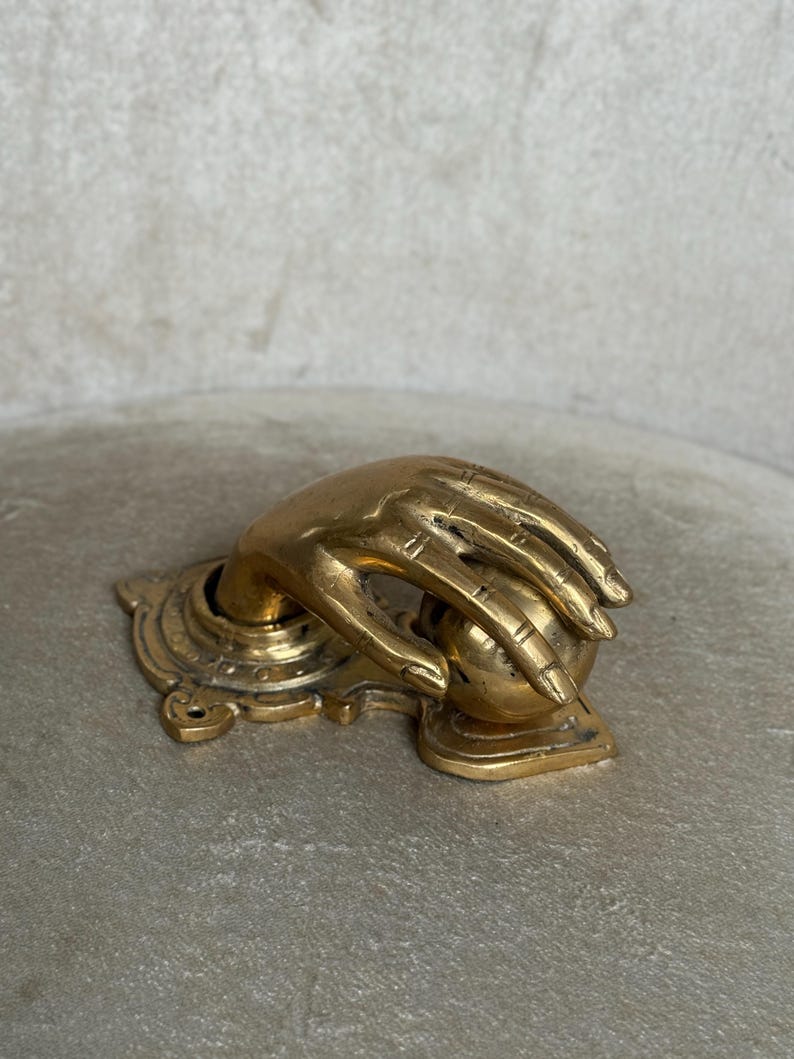 May include: A brass door knocker in the shape of a hand grasping a sphere. The hand is detailed with visible knuckles and lines, mounted on an ornate base. The finish is a warm, aged gold.