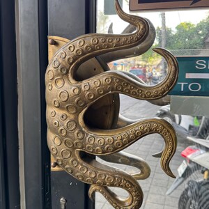 May include: A brass-coloured door handle in the shape of an octopus, featuring detailed suckers and tentacles. The handle is fixed to a black door, with a keyhole below. The design is elaborate and striking.