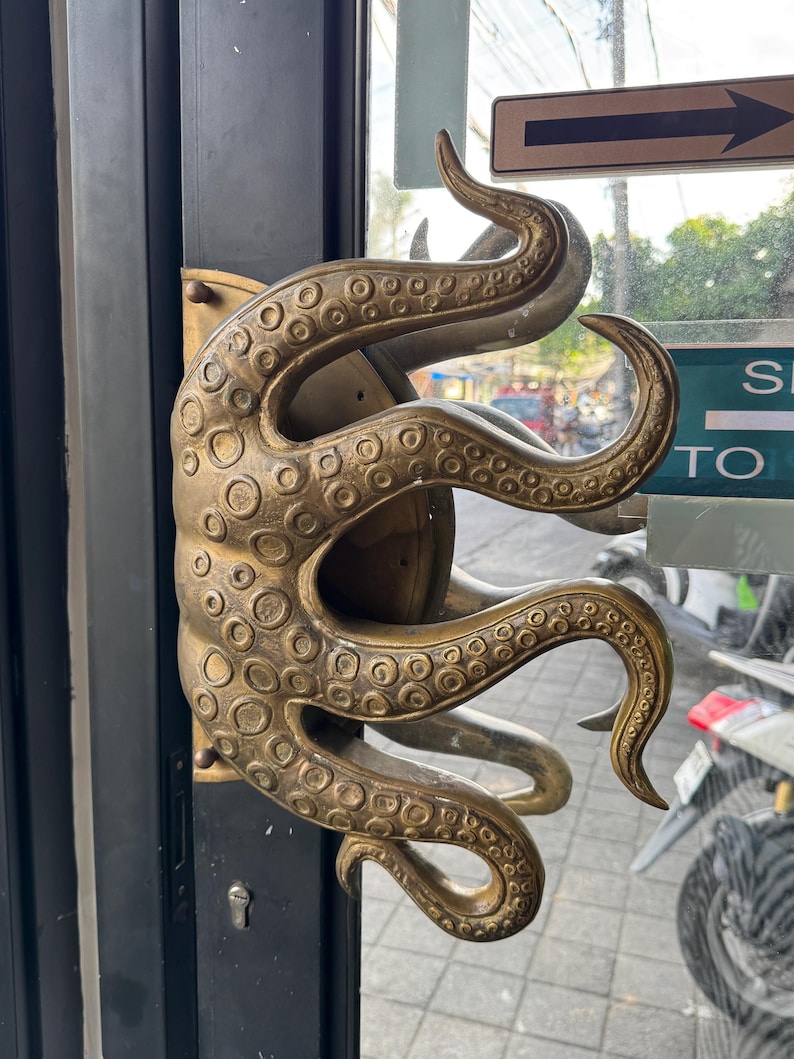 May include: A brass-coloured door handle in the shape of an octopus, featuring detailed suckers and tentacles. The handle is fixed to a black door, with a keyhole below. The design is elaborate and striking.