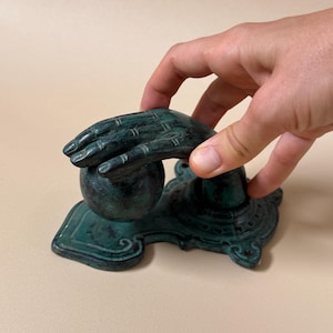 May include: A teal-colored decorative hand sculpture with a ball and ornate base. The sculpture has a weathered appearance, with visible texture and detail. The hand is positioned as if holding the ball. The base has a detailed, decorative design.