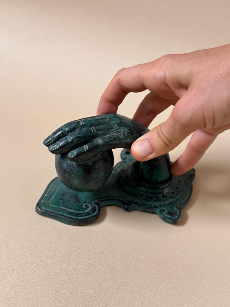 May include: A teal-colored decorative hand sculpture with a ball and ornate base. The sculpture has a weathered appearance, with visible texture and detail. The hand is positioned as if holding the ball. The base has a detailed, decorative design.