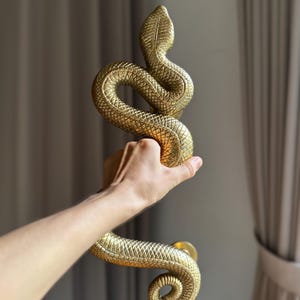 May include: A golden door handle in the shape of a coiled snake. The handle is held in a hand, showcasing the intricate scale details and the serpent's head. The handle is made of metal and has a shiny finish.