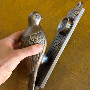 May include: A brass door knocker featuring a bird figure attached to a rectangular backplate. The bird has detailed wing feathers and a smooth head. The backplate displays a circular strike plate and embossed linear patterns on its surface.