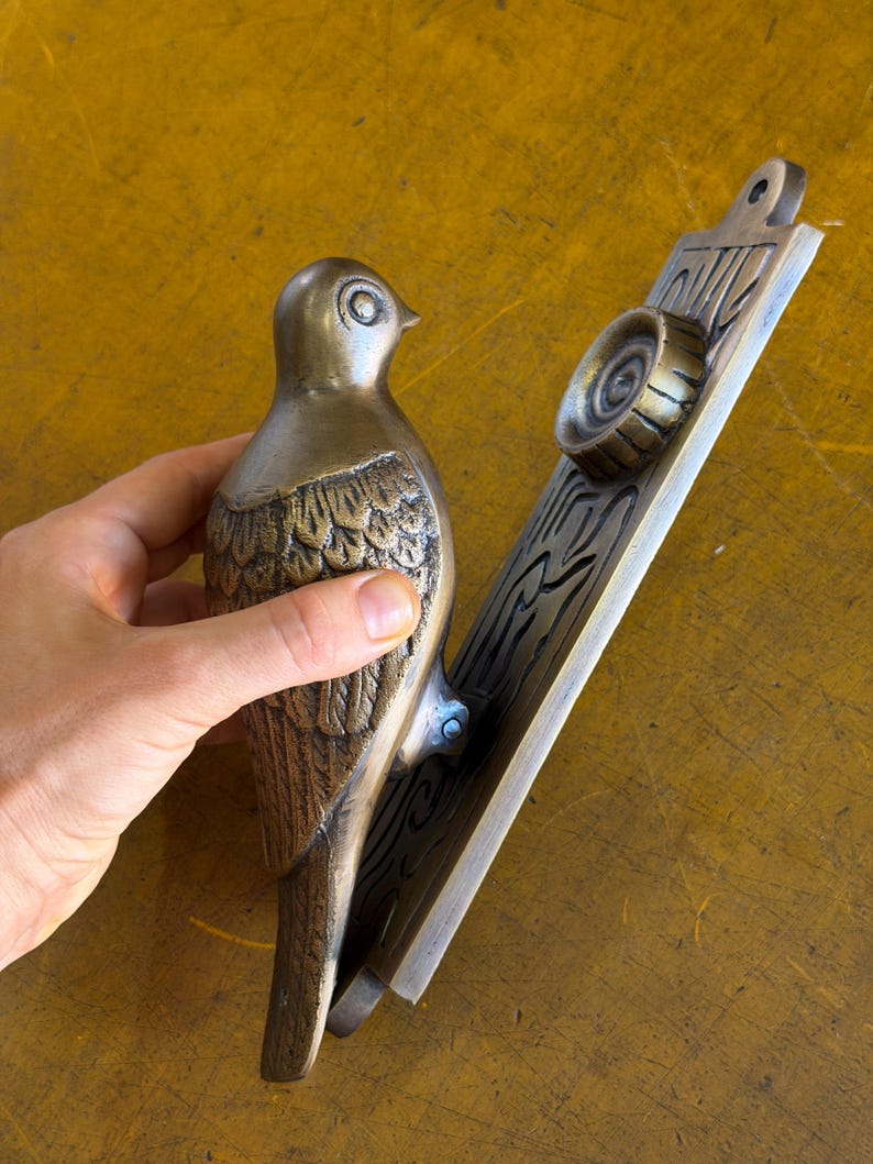 May include: A brass door knocker featuring a bird figure attached to a rectangular backplate. The bird has detailed wing feathers and a smooth head. The backplate displays a circular strike plate and embossed linear patterns on its surface.