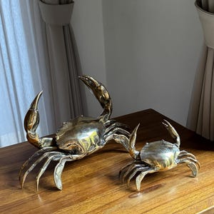 May include: Two brass crab sculptures with detailed claws and bodies sit on a wooden surface. The larger crab has its claws raised, while the smaller one has its claws lowered. The crabs are a decorative item.