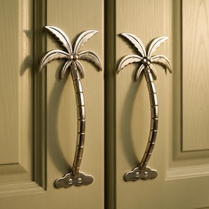May include: Two silver-toned palm tree-shaped cabinet handles mounted on a light green door. The handles have a curved trunk design and a detailed palm frond top. The door has a recessed panel design.