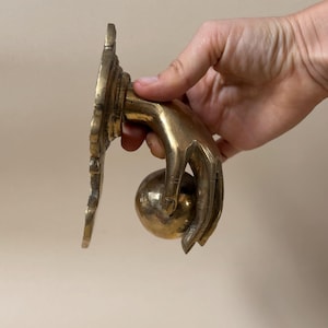 May include: A brass door knocker in the shape of a hand holding a sphere. The knocker has a decorative backplate with an ornate design. The hand and sphere are polished to a shine, and the overall design is vintage.