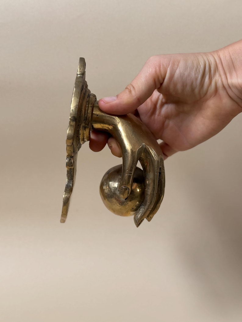 May include: A brass door knocker in the shape of a hand holding a sphere. The knocker has a decorative backplate with an ornate design. The hand and sphere are polished to a shine, and the overall design is vintage.