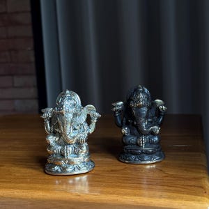 May include: Two small, ornate statues of the Hindu deity Ganesha. The statue on the left is silver, while the statue on the right is black. Both statues are sitting on a wooden surface.