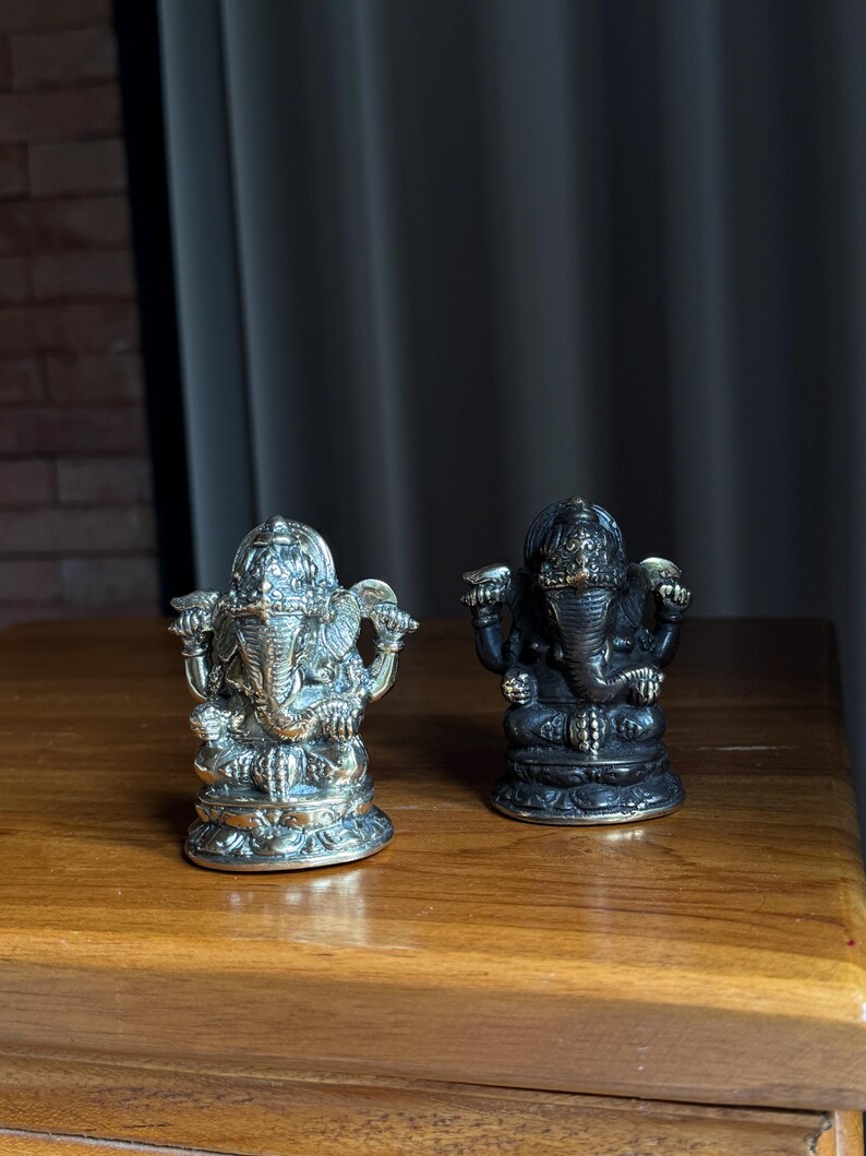 May include: Two small, ornate statues of the Hindu deity Ganesha. The statue on the left is silver, while the statue on the right is black. Both statues are sitting on a wooden surface.