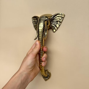 May include: A gold-colored elephant-shaped door handle. The handle is held against a neutral-colored wall. The elephant head features detailed ears and a trunk, with a curved handle for gripping. The handle is made of metal.