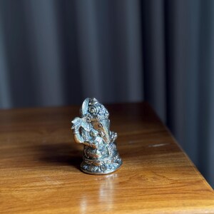 May include: A small, silver-colored metal statue of Ganesha, the Hindu god of wisdom and prosperity, sitting on a wooden surface.