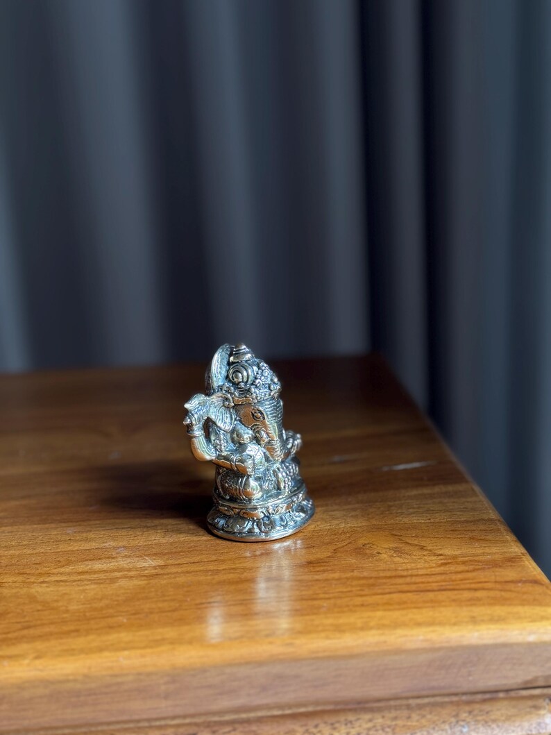 May include: A small, silver-colored metal statue of Ganesha, the Hindu god of wisdom and prosperity, sitting on a wooden surface.