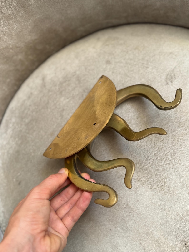 May include: A vintage brass wall-mounted coat rack. It features a half-circle backplate and four curved hooks. The rack has a warm, golden hue and a unique, stylized design. The hooks are curved and resemble stylized tentacles.