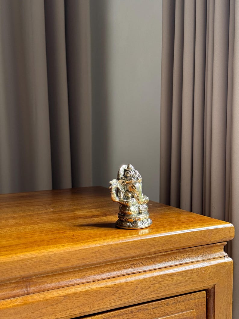 May include: A silver Ganesha statue with intricate details sits on a wooden table. The statue is about 5 inches tall and has a trunk pointing to the right.