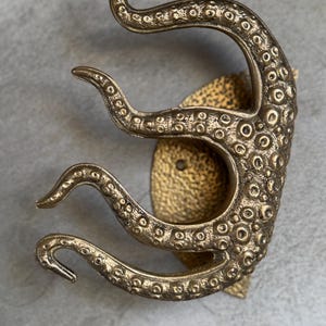 May include: A bronze-coloured octopus tentacle wall hook. The hook features detailed suckers and a textured surface. The design is mounted on a textured, gold-coloured base, creating a unique nautical or fantasy-themed decor item.