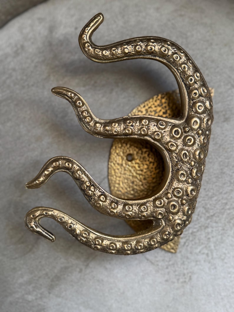 May include: A bronze-coloured octopus tentacle wall hook. The hook features detailed suckers and a textured surface. The design is mounted on a textured, gold-coloured base, creating a unique nautical or fantasy-themed decor item.