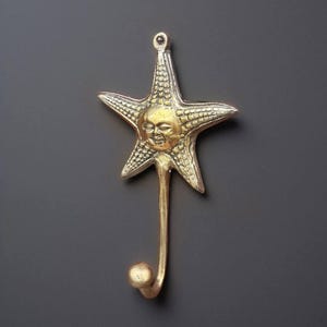 Puede incluir: Un colgador de pared de color latón con forma de estrella de mar con una cara de sol sonriente. El colgador tiene un tallo curvo y una pequeña bola en el extremo. La estrella de mar mide aproximadamente 10 cm de ancho.
