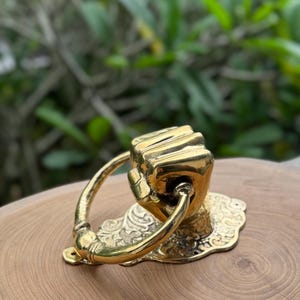 May include: A brass door knocker in the shape of a clenched fist. The knocker has a curved handle and is mounted on a decorative, ornate base. The finish is a shiny gold color.