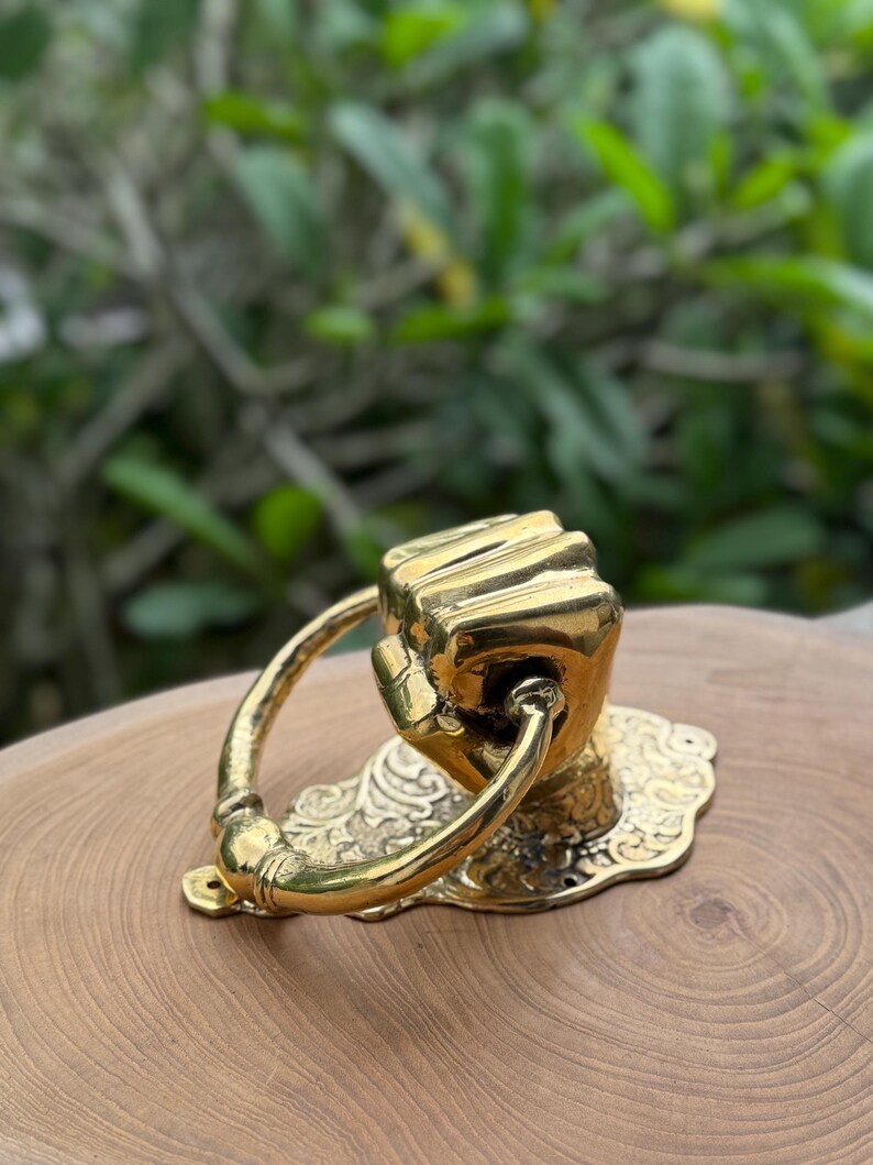 May include: A brass door knocker in the shape of a clenched fist. The knocker has a curved handle and is mounted on a decorative, ornate base. The finish is a shiny gold color.