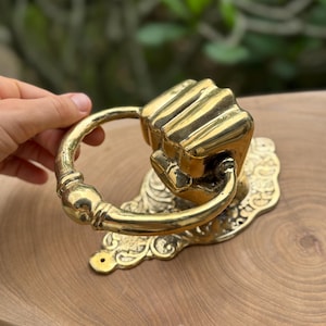 May include: A brass door knocker shaped like a clenched fist. The knocker has a ring handle and is mounted on an ornate, decorative backplate. The finish is shiny gold.