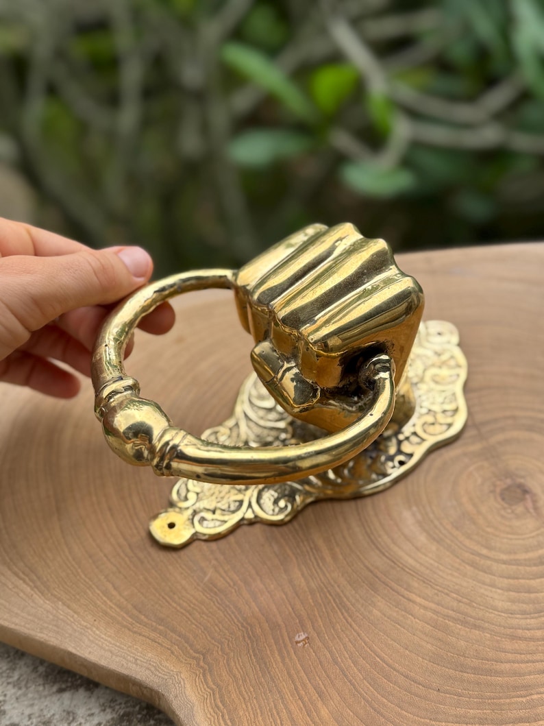 May include: A brass door knocker shaped like a clenched fist. The knocker has a ring handle and is mounted on an ornate, decorative backplate. The finish is shiny gold.