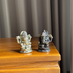 May include: Two small, ornate, gold and bronze Ganesha statues on a wooden surface. The statues are facing forward and have intricate details.