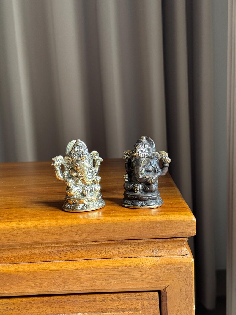 May include: Two small, ornate, gold and bronze Ganesha statues on a wooden surface. The statues are facing forward and have intricate details.