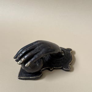 May include: A dark bronze-colored sculpture of a hand cradling a sphere. The fingers are slightly curved, with gold-colored accents on the fingertips. The hand rests on a decorative base with ornate detailing.