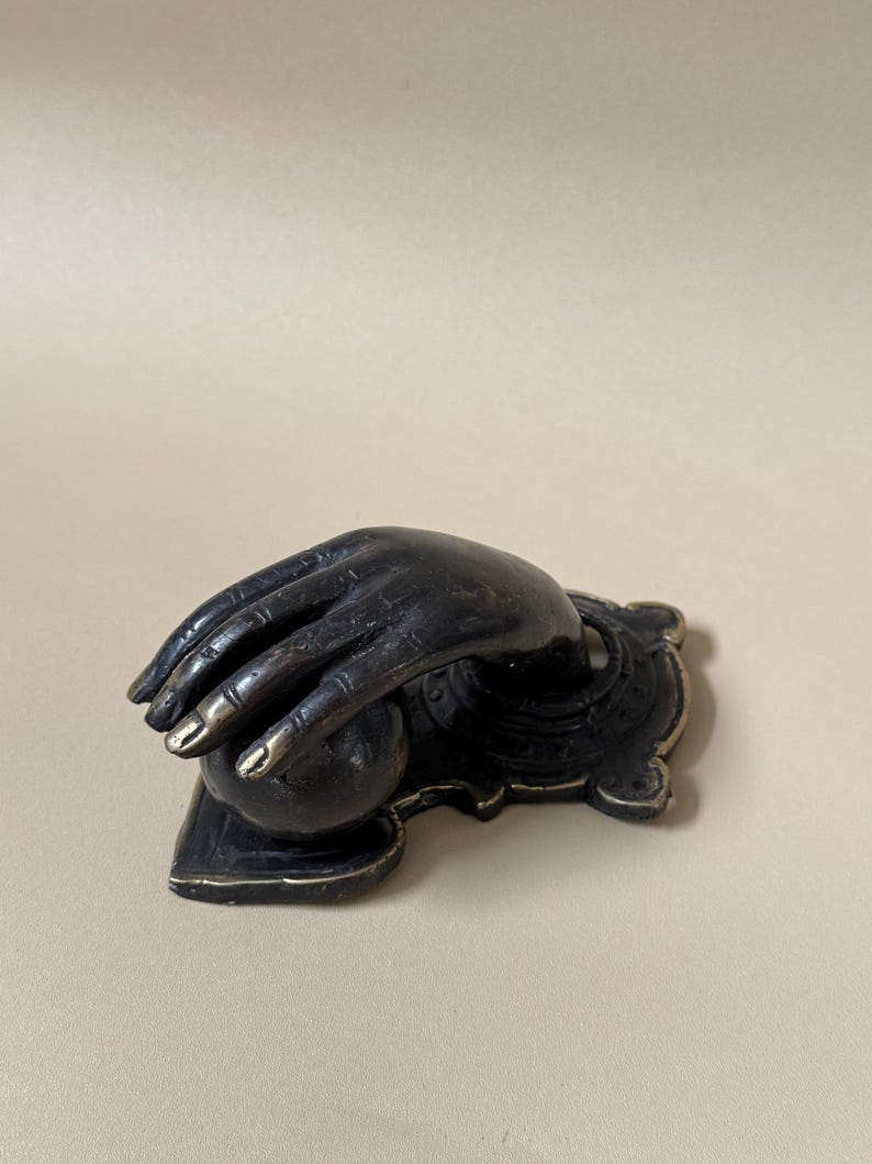 May include: A dark bronze-colored sculpture of a hand cradling a sphere. The fingers are slightly curved, with gold-colored accents on the fingertips. The hand rests on a decorative base with ornate detailing.