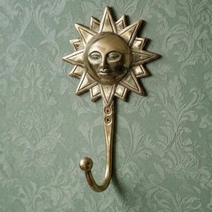 Könnte beinhalten: Ein Wandhaken aus Messing mit einem Sonnengesicht-Design und strahlenförmigen, dreieckigen Spitzen. Der Haken hat einen geschwungenen Arm, der in einer kleinen Kugel endet, und ist mit zwei sichtbaren Schrauben unter dem Gesicht befestigt.