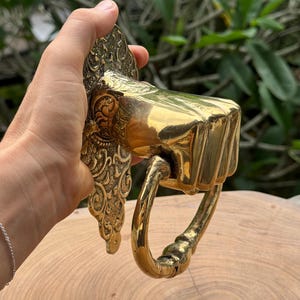 May include: Ornate brass door knocker with a rectangular striking plate and a curved handle. The knocker features detailed scrollwork and a polished, reflective surface, showcasing a vintage aesthetic.