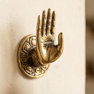 Op de afbeelding: Een messing wandhaak in de vorm van een open hand met de vingers omhoog gericht. De hand is bevestigd aan een ronde basis met een decoratief bloem- of bloembladachtig reliëfpatroon. Het metaal heeft een verweerde, antieke afwerking.