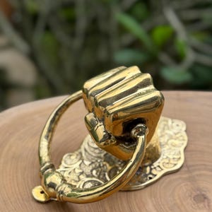 May include: A brass door knocker featuring a fist design. The knocker has a ring handle and is mounted on a decorative base with ornate detailing. The finish is a shiny gold color, and the design is highly detailed.