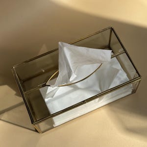 May include: A rectangular tissue box with a clear glass body and a gold-colored metal frame. White tissues are visible inside, with one tissue partially pulled out. The box is on a neutral-colored surface.