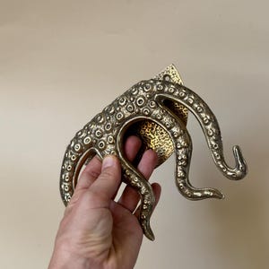 May include: A bronze-coloured octopus-shaped door handle. The handle has a textured surface with small circular details and curved tentacles. The handle is attached to a rectangular plate with a hammered finish.