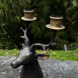 May include: A black metal deer head candle holder with two gold candle cups. The deer head has a detailed design and the candle cups have a geometric pattern.