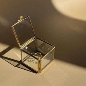 Op de afbeelding: Een kleine, open glazen doos met goudkleurige metalen randen. Een gouden ring ligt in de doos. De doos staat op een beige oppervlak, met een schaduw.
