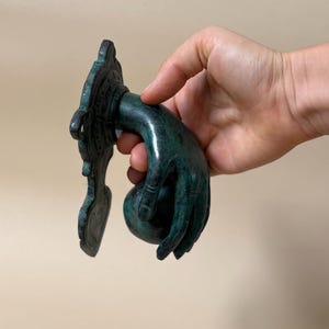 May include: A detailed, verdigris-colored door knocker in the shape of a hand. The hand is holding a round ball, and the knocker has an ornate, decorative backplate. The finish has a green patina.