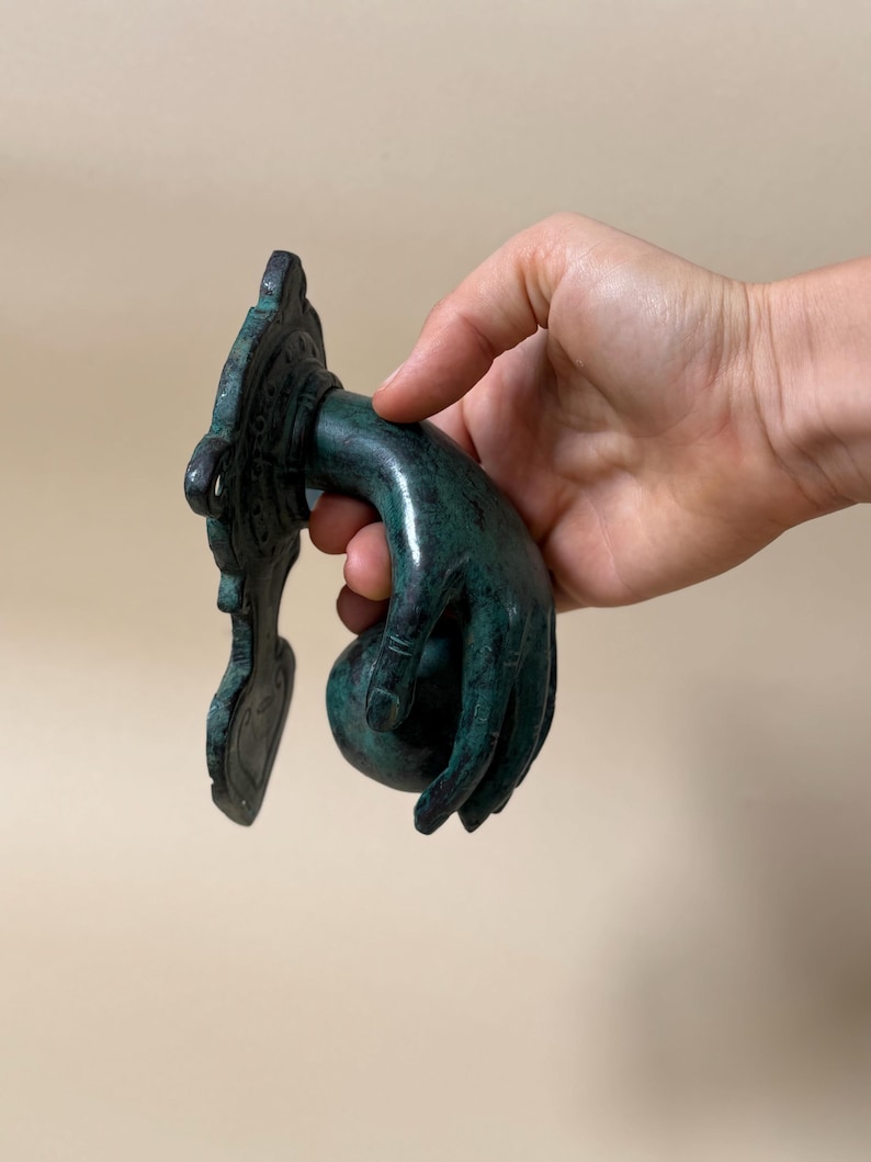 May include: A detailed, verdigris-colored door knocker in the shape of a hand. The hand is holding a round ball, and the knocker has an ornate, decorative backplate. The finish has a green patina.