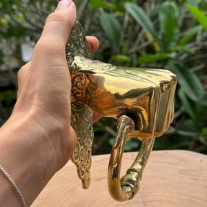 May include: A brass door knocker with a fist design. The knocker is held in a hand and features intricate detailing on the backplate and a curved handle. The finish is a shiny gold color, reflecting light.