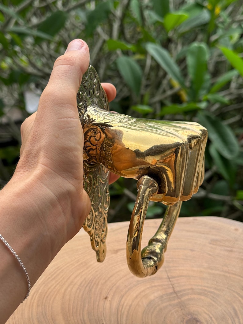 May include: A brass door knocker with a fist design. The knocker is held in a hand and features intricate detailing on the backplate and a curved handle. The finish is a shiny gold color, reflecting light.
