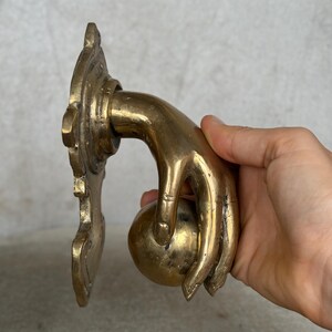 May include: A brass door knocker in the shape of a hand holding a sphere. The hand is detailed with visible knuckles and a slight curve. The knocker is mounted on a decorative backplate with ornate edges.