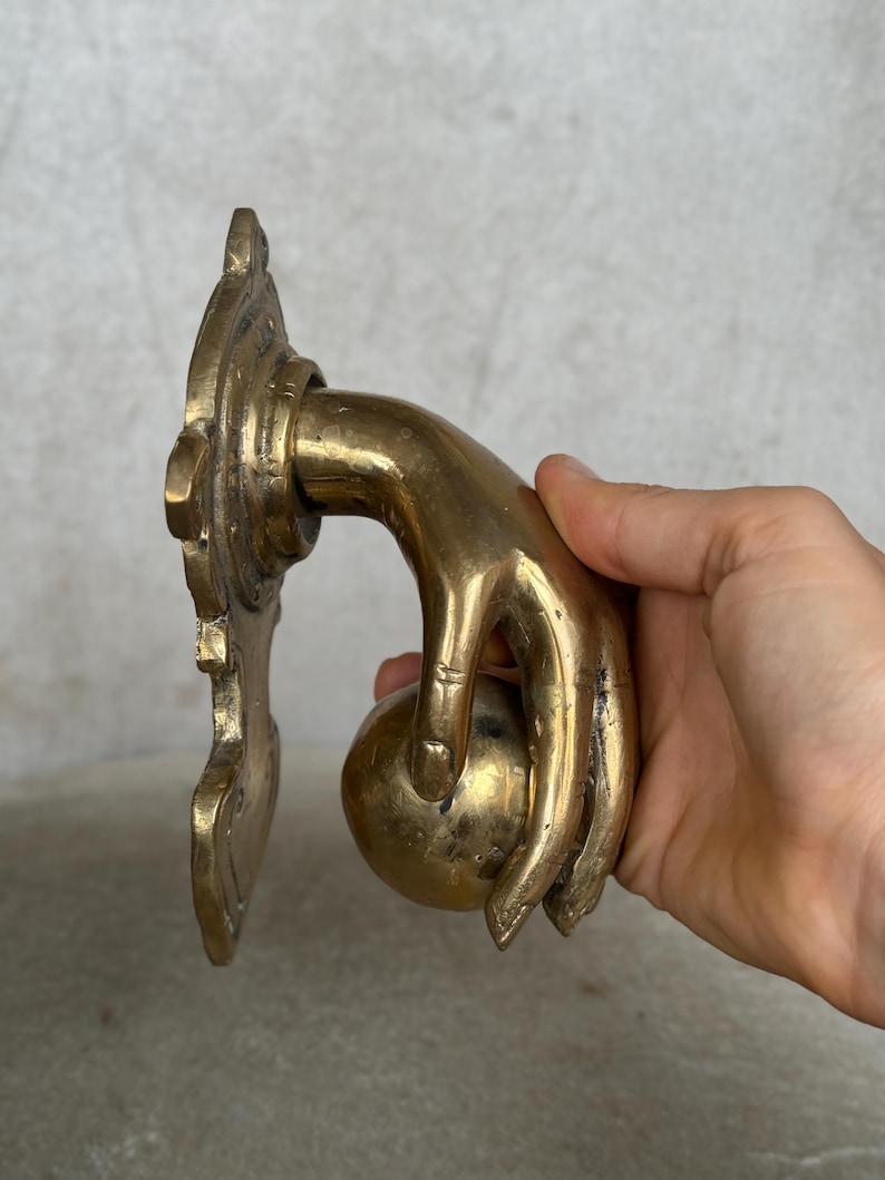 May include: A brass door knocker in the shape of a hand holding a sphere. The hand is detailed with visible knuckles and a slight curve. The knocker is mounted on a decorative backplate with ornate edges.