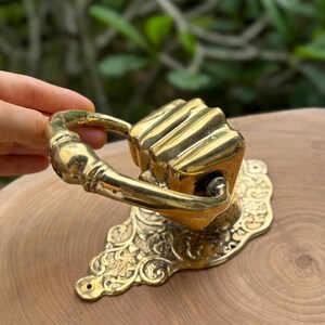 May include: A brass door knocker with a decorative floral base and a handle. The knocker has a rectangular shape with vertical lines. The handle is curved and attached to the rectangular knocker.