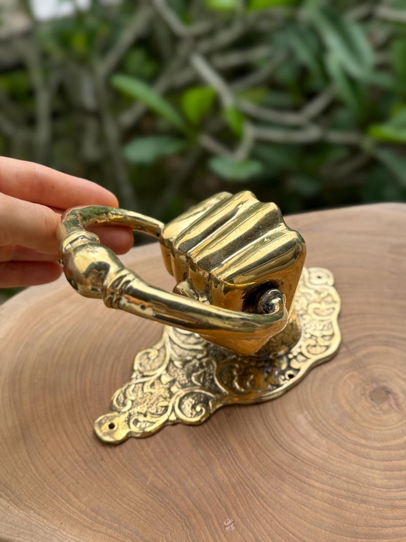 May include: A brass door knocker with a decorative floral base and a handle. The knocker has a rectangular shape with vertical lines. The handle is curved and attached to the rectangular knocker.