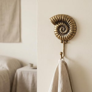 May include: A brass-colored wall hook in the shape of a nautilus shell. The hook is mounted on a cream-colored wall and holds a beige towel. The nautilus shell design adds a touch of coastal decor.