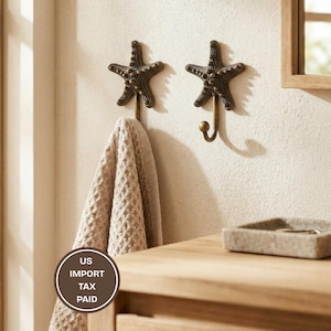 May include: Two bronze starfish-shaped wall hooks with a towel hanging from one. The hooks are mounted on a light-colored wall. A wooden cabinet and a small stone tray are visible in the foreground. The hooks add a coastal theme to any room.