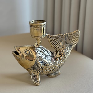 May include: A brass-colored candle holder shaped like a fish. The fish has detailed scales, fins, and an open mouth. A small cup-shaped holder sits on the fish's back to hold a candle. The candle holder is a decorative item.