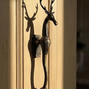 May include: A decorative door handle in the shape of a deer. The handle is made of dark bronze metal and features intricate details, including the deer's antlers and a textured body. The handle is mounted on a rectangular plate.